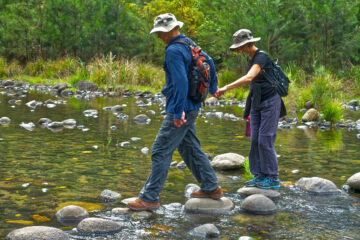 Ein Mann und eine Frau halten sich an der Hand und überqueren gemeinsam einen Bach. Foto: pixabay.com / MemoryCatcher