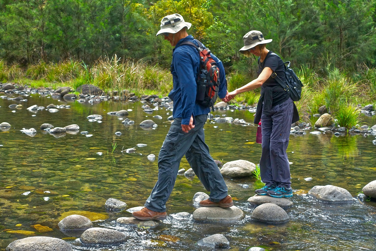 Ein Mann und eine Frau halten sich an der Hand und überqueren gemeinsam einen Bach. Foto: pixabay.com / MemoryCatcher